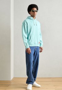 Felpa verde menta con cappuccio e cordoncino, tasca frontale a marsupio e una toppa con logo. Abbinata a jeans blu e sneakers bianche.