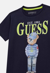 T-shirt blu navy con grafica di un orso che indossa un maglione a righe e un cappello. Il design include scritte audaci in bianco e verde e jeans strappati.