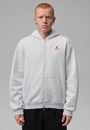 Młody mężczyzna z krótkimi czerwonymi włosami, noszący srebrne kolczyki koła oraz jasnoszary bluza z kapturem na zamek z czerwonym logo Air Jordan na piersi.