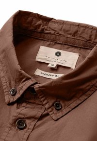 Camisa marrón de botones con cuello, que presenta botones negros y un cuello interior texturizado. Las etiquetas incluyen información sobre la marca y la talla.
