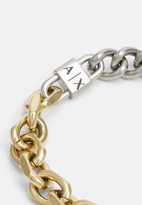 Metallkedjearmband med växelvis gyllene och silverlänkar, med en rektangulär silverlåsningsanordning graverad med "A/X." Slät, polerad yta.