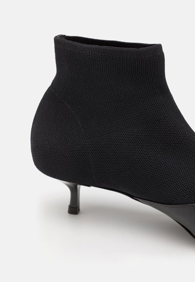 Sorte ankelsko med en struktureret strikoverdel, spidse tæer og en tynd, stiletto hæl. Materialet er strækbart og tætsiddende.