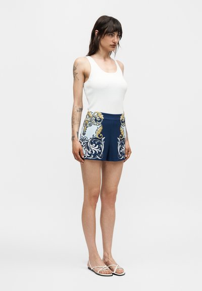 Γυναίκα που στέκεται, φοράει λευκό αμάνικο τοπ, σκούρο μπλε σορτς με περίτεχνα χρυσά και λευκά σχέδια και λευκά σανδάλια με λουριά.