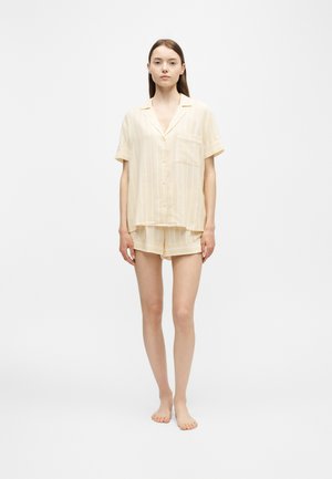 Jeune femme debout pieds nus portant un haut à manches courtes boutonné rayé jaune pâle et un short assorti sur fond blanc.