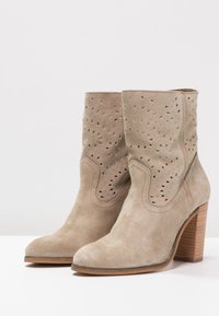 Botas de tobillo de ante beige con patrones cortados a láser, punteras redondeadas y tacones de madera apilados. Parte superior texturizada con acabado suave.