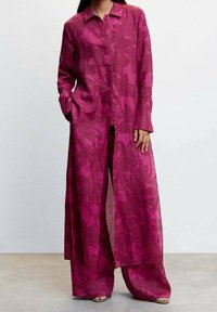 Kvinde iført en lang, løstsiddende magenta blomstret skjortekjole med matchende vide bukser og sandaler, stående med den ene hånd i lommen.