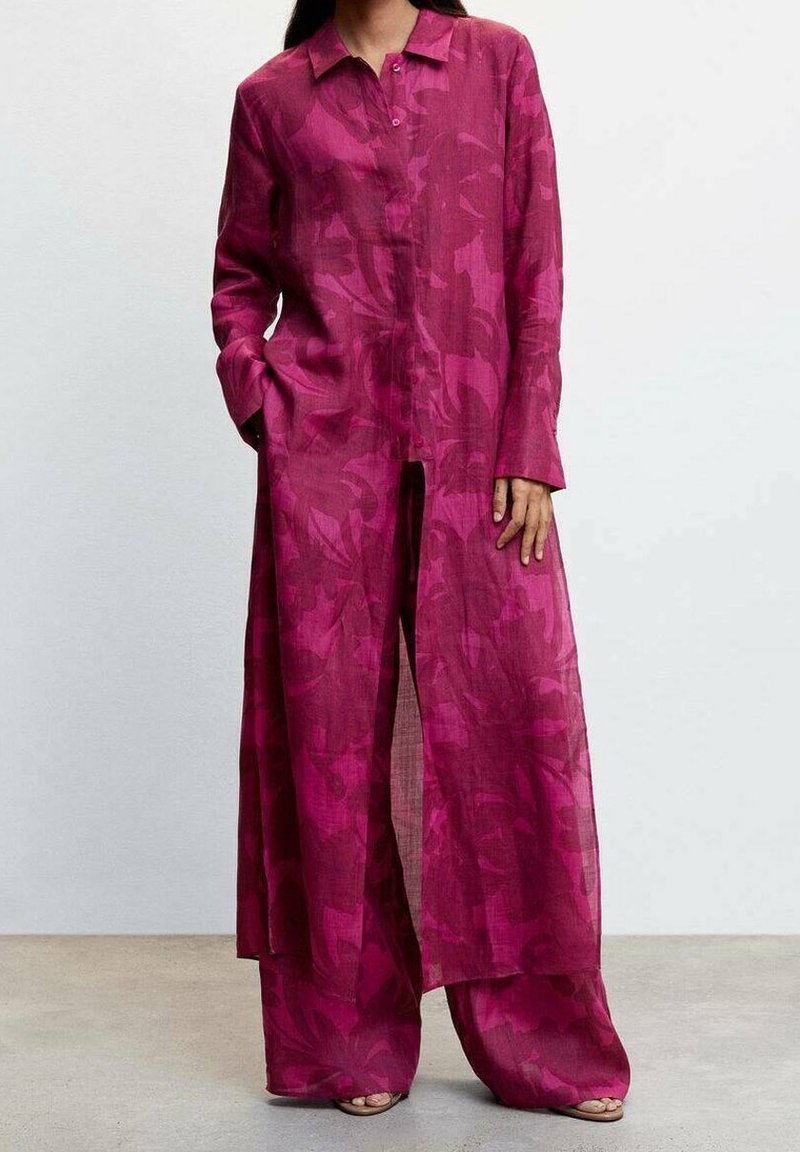 Kvinde iført en lang, løstsiddende magenta blomstret skjortekjole med matchende vide bukser og sandaler, stående med den ene hånd i lommen.