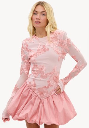 Femme blonde portant une robe rose à manches longues avec broderies florales et une jupe volumineuse, posant avec une main sur la hanche.