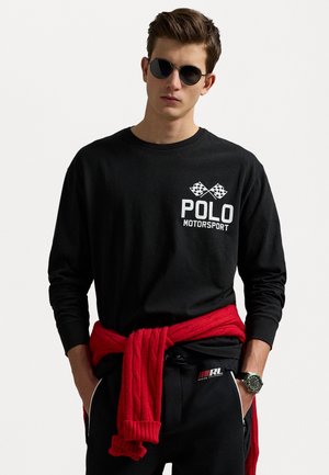 Schwarzes langärmliges T-Shirt mit weißem 'POLO MOTORSPORT'-Logo und karierten Flaggen. Roter Strickpullover um die Taille gebunden. Schwarze Hose.