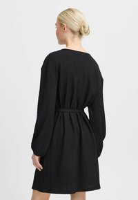 Robe noire en tissu texturé, avec des manches longues, une coupe loose et une ceinture à nouer à la taille. L'encolure est ronde.