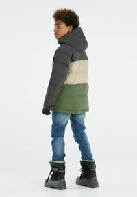 Veste matelassée avec capuche en gris, beige et vert olive. Dotée de poignets élastiques et de contrastes noirs. Portée avec un jean bleu et des bottes sombres.
