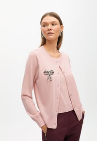 Rosa stickad kofta med långa ärmar, rund halsringning och pärlknappar. Har en dekorativ brosch formad som en rosett gjord av rhinestones.