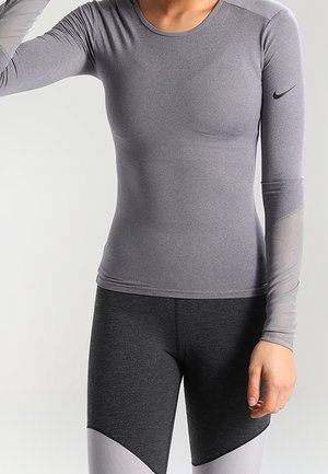Personne portant un haut de sport gris à manches longues ajusté et un legging gris foncé avec des panneaux gris clair, debout devant un fond uni.