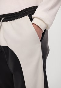 Personne portant un pantalon noir et blanc avec une taille élastique et une main dans la poche avant, vêtue d'un haut blanc à manches longues.