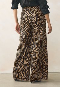 Pantalones anchos de pierna ancha en tonos marrón y dorado con un patrón de rayas de tigre, con cintura alta y textura suave, combinados con una camiseta y zapatos negros.