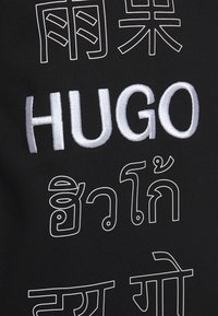 Tela negra con texto blanco bordado, que incluye "HUGO" y varios caracteres en diferentes idiomas, con contornos contrastantes.