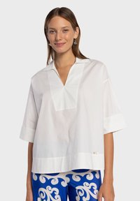 Blouse en coton blanc avec un col en V, des manches courtes et un ourlet droit. Présente une légère brillance et une petite étiquette décorative en bas.