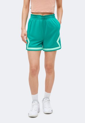 Persona con pantalones cortos deportivos turquesa con ribete blanco, un top peach corto, calcetines blancos y zapatillas deportivas blancas, de pie con una mano en el bolsillo.