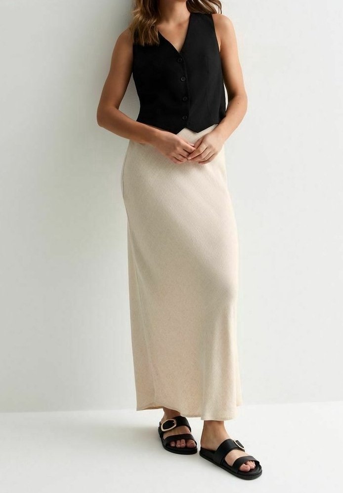 Femme portant un gilet noir sans manches boutonné, une jupe beige longue jusqu’aux chevilles et des sandales noires avec des détails de boucle métallique.