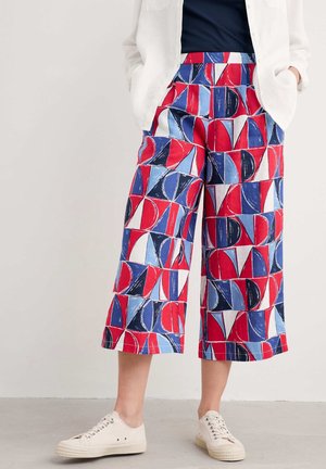 Osoba nosząca białe trampki, spodnie culotte z abstrakcyjnym geometrycznym wzorem w czerwonym, niebieskim i białym kolorze, granatową bluzkę i białą kurtkę z rękami w kieszeniach.