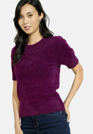 Maglione corto a maniche corte di colore viola, realizzato in un materiale morbido e peloso, con collo rotondo e vestibilità aderente in vita. Indossato con jeans scuri.