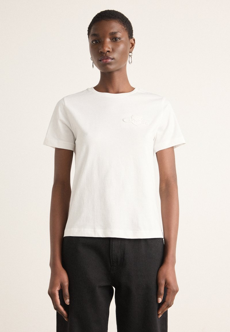 Jeune femme aux cheveux courts portant un T-shirt blanc uni à manches courtes et un pantalon noir, debout devant un fond clair.