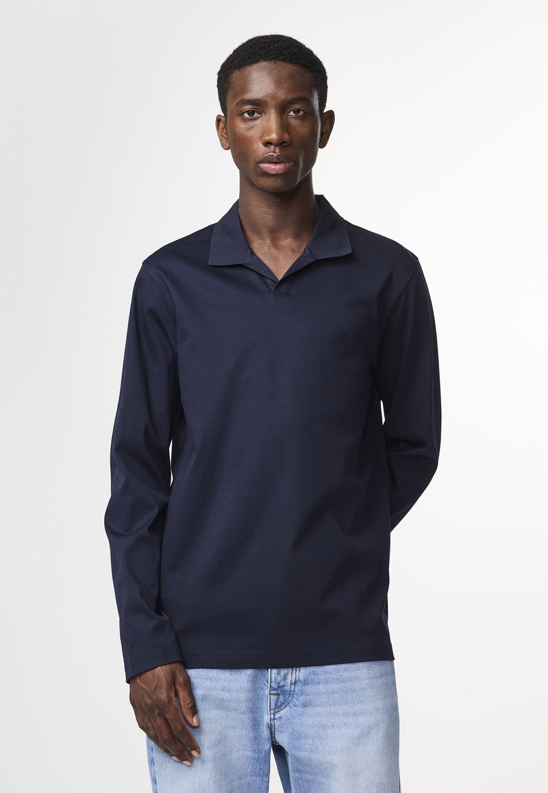 Polo à manches longues navy avec col plat, fabriqué en tissu doux. Présente une coupe décontractée et des détails de couture subtils. Associé à un jean bleu clair.