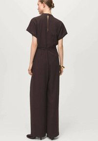 Brązowy kombinezon z krótkimi rękawami, zapięciem na plecach w kształcie klucza, szerokimi nogawkami i paskiem w talii. Gładka tekstura, minimalistyczny design.