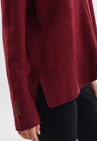 Burgundowy sweter z prążkowanym wykończeniem oraz mankietami, bocznymi rozcięciami i metką z tkaniny. Miękka struktura z luźnym krojem.