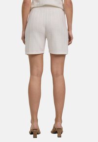 Shorts en maille blanche avec des côtes verticales, une taille élastique et des ourlets. Portés avec des talons marron, affichés de dos.