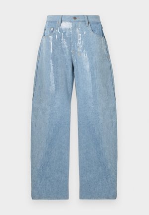 Nainen pukeutuneena vaaleansiniseen, paljettikuviolliseen lyhyeen takkiin ja siihen sopiviin leveälahkeisiin housuihin, valkoinen toppi alla ja mustat korolliset sandaalit.