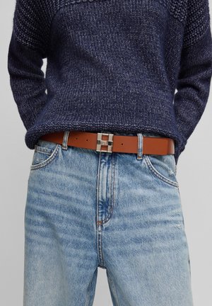 Personne portant un pull tricoté bleu foncé rentré dans un jean taille haute bleu clair avec une ceinture en cuir marron et une boucle argentée.