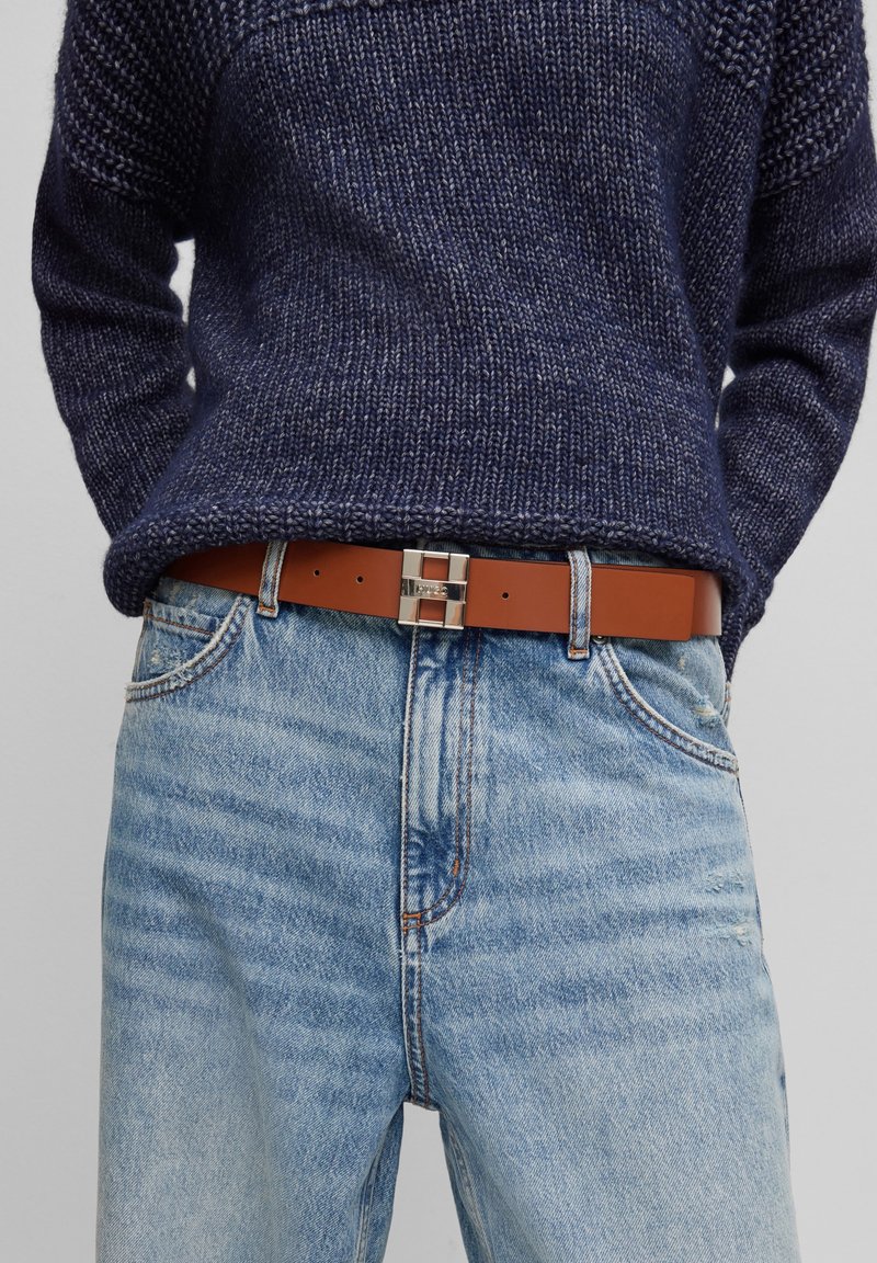 Personne portant un pull tricoté bleu foncé rentré dans un jean taille haute bleu clair avec une ceinture en cuir marron et une boucle argentée.