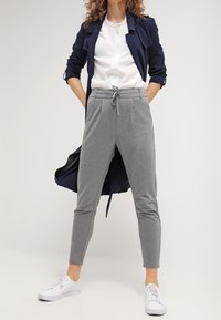 Pantalones gris ajustados con cintura con cordón, combinados con una blusa blanca y un abrigo trinchera azul marino. Llevados con zapatillas blancas. Textura suave.