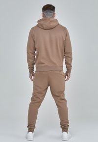 Brown hoodie and matching joggers made of soft fabric. Features a hood, ribbed cuffs, and a small pocket detail on the back. White sneakers.