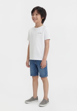 Usmievajúci sa chlapec v bielom tričku s krátkym rukávom značky Calvin Klein, modrých riflových šortkách a šedých slip-on topánkach stojaci pred bielym pozadím.