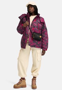 Paarse en zwarte bloemen pufferjack, crèmekleurige joggingsbroek, tan laarzen met roze veters en een zwarte schoudertas. Getextureerde materialen overal.