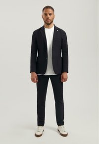 Sacou închis la culoare cu două buzunare frontale, purtat peste un tricou alb, asortat cu pantaloni închiși la culoare și adidași albi. Material neted, croială ajustată.