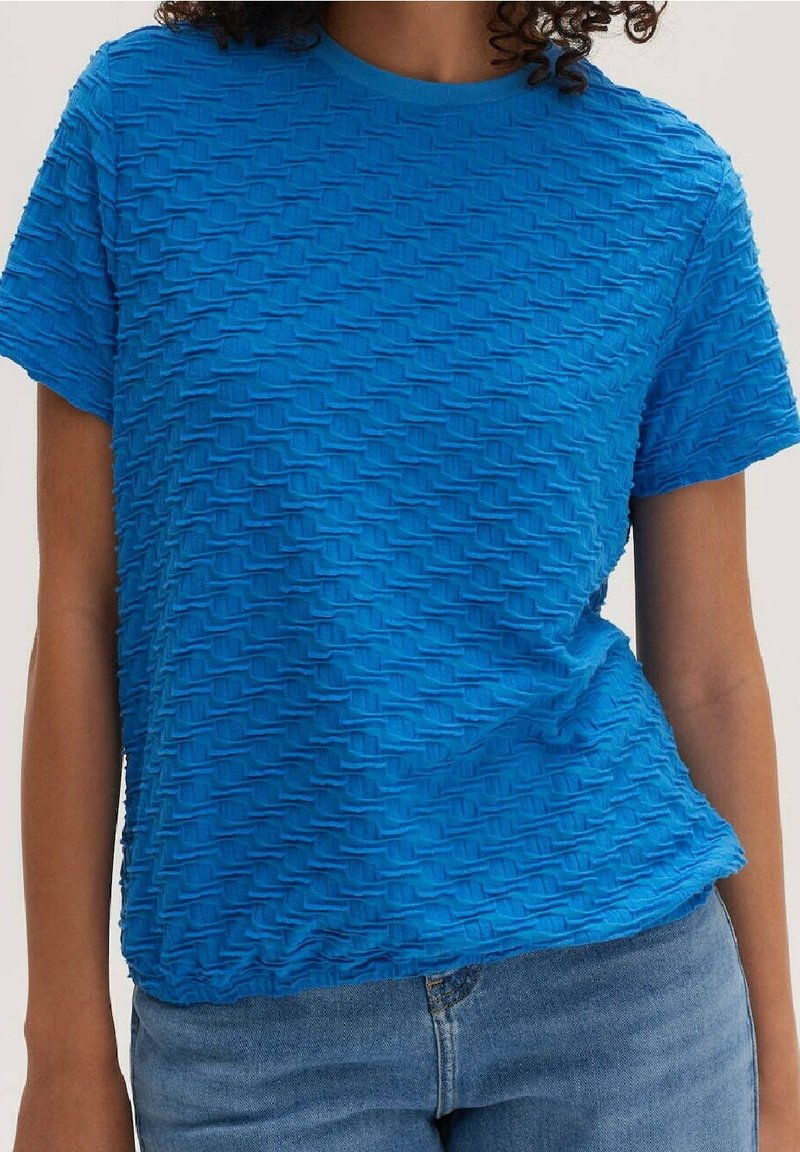 Personne portant un haut à manches courtes texturé bleu vif associé à un jean en denim bleu clair, coupé au niveau du torse.
