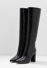 Sorte knæhøje støvler med en blank krokodilletekstur, spids tå og blokhæle. Fremstillet af syntetisk materiale.