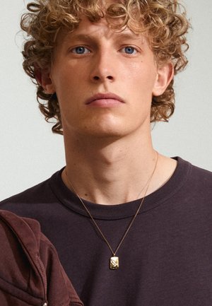 Collar de péndulo de oro con un charm rectangular texturizado, llevado sobre una cadena delgada, encima de un sudadero de cuello redondo de color púrpura oscuro.