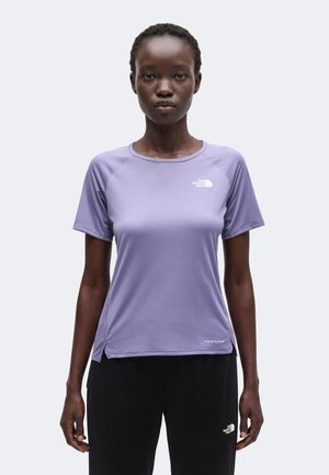 Γυναίκα που φοράει στενή μοβ μπλούζα North Face με κοντά μανίκια και μαύρο παντελόνι North Face, στέκεται μπροστά σε απλό ανοιχτό φόντο.