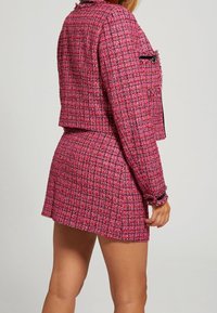 Vrouw in een rode en roze gehaakte jas met een bijpassende hooggesneden mini-jurk, staande tegen een effen achtergrond.