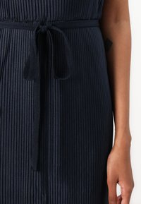 Abito plissettato di un blu navy con cintura annodata in vita, caratterizzato da una texture liscia e un motivo a righe verticali. Mano visibile sul lato destro.