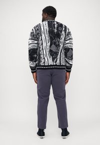 Black and white patterned bomber jacket with textured design; ribbed cuffs and hem. Worn with gray pants and black sneakers.