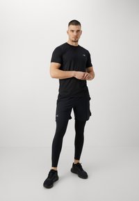 Camiseta deportiva negra, diseño ajustado, de mangas cortas. Combinada con pantalones cortos negros, con una capa de mallas negras. Las zapatillas negras completan el look.