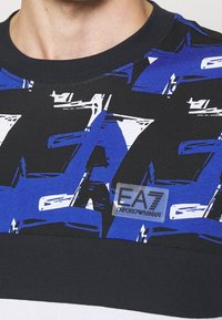 Tílko s krátkým rukávem a kulatým výstřihem v černé, bílé a modré barvě, zdobené výrazným abstraktním vzorem a logem s nápisem "EA7 Emporio Armani."