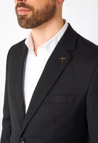 Blazer negro con un acabado satinado, cierre de un solo botón y un pequeño acento cruzado naranja en la solapa, llevado sobre una camisa blanca.
