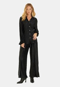 Conjunto de dos piezas de satén negro que incluye una camisa con botones, de manga larga y puños con volantes, combinado con pantalones de pierna ancha. Tela texturizada.