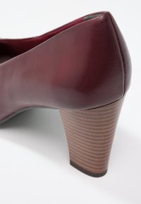 Chaussure à talon haut en cuir bordeaux avec un talon en bois strié et texturé. Surface lisse avec un bout rond et des lignes épurées.